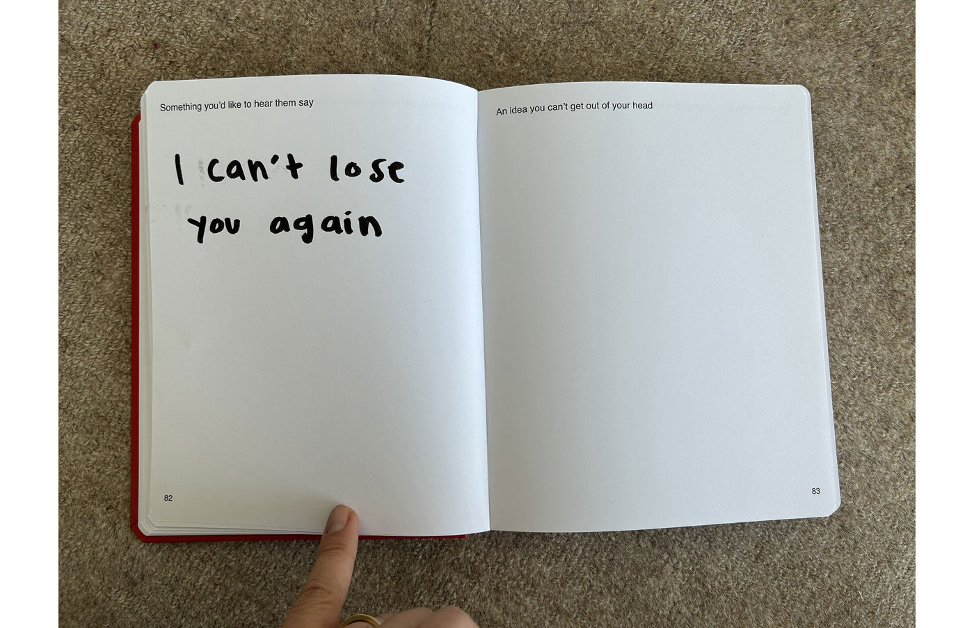 We're Not Really Strangers Journal. Front facing view of someone holding down one of the journal pages with prompt "Something you'd like to hear them say" with response in black marker reading "I can't lose you again." A second prompt also showing on blank page reading "An idea you can't get out of your head."